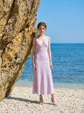 Woman in a lilac midi dress with a V-neckline and sleeveless design, posing by a rock on a beach with the ocean in the background.