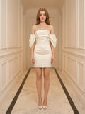 Woman in a cream off-the-shoulder satin mini dress with puff sleeves, standing in a hallway.