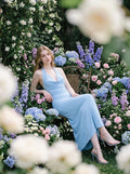 Woman sitting in a light blue maxi halter dress surrounded by a colorful floral garden.
