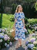 Woman wearing a white and blue floral print midi dress with puff sleeves and a cut-out waist, walking in a garden.