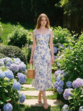Woman wearing a white midi dress with a blue floral print and tie shoulder straps, walking in a garden with hydrangeas.
