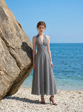 Woman wearing a black and white gingham check halter neck maxi dress with a plunging V-neck and a defined empire waist, posing on a beach with large rocks and the ocean in the background.