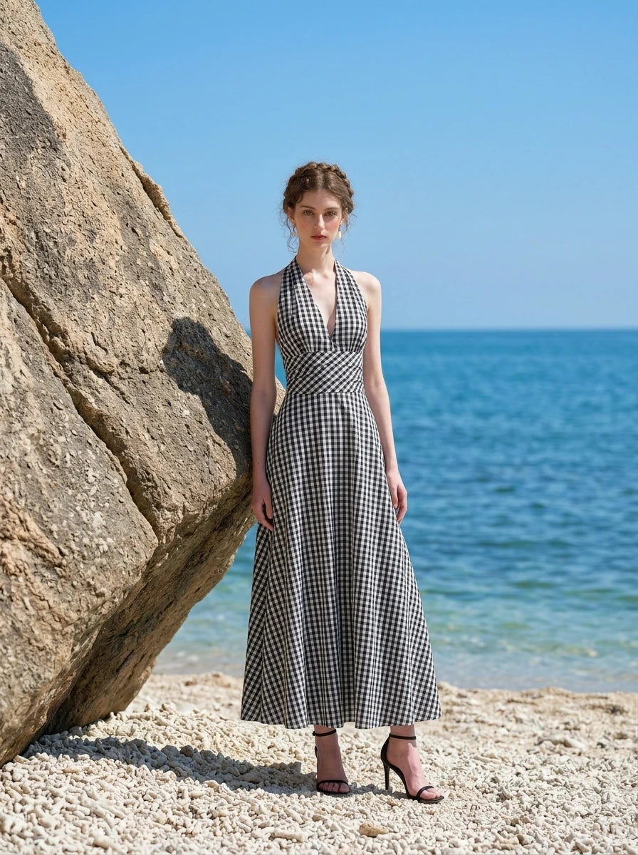 Woman wearing a black and white gingham check halter neck maxi dress with a plunging V-neck and a defined empire waist, posing on a beach with large rocks and the ocean in the background.