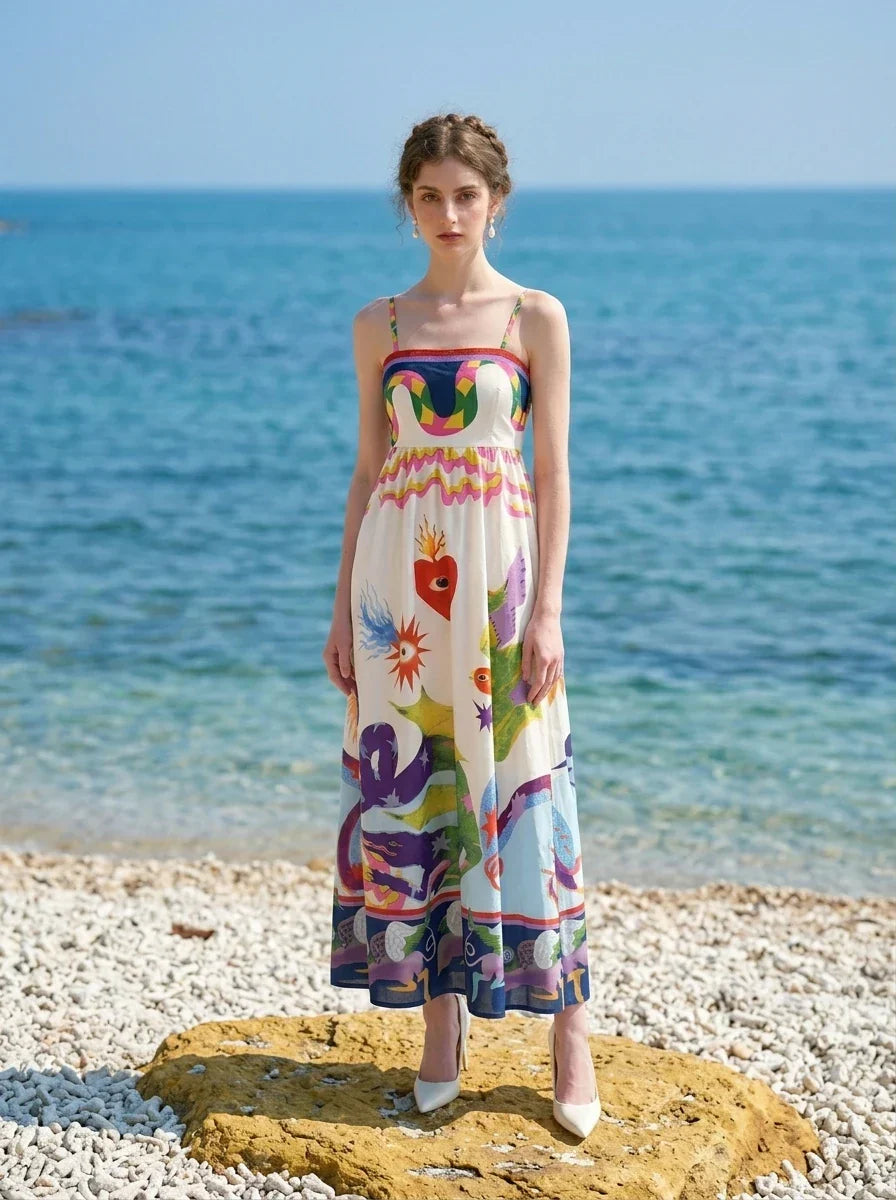Woman wearing a white midi dress with a colorful graphic print and spaghetti straps standing on a beach with the ocean in the background