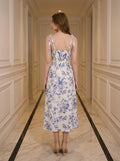 Back view of a woman in a floral print midi dress with tie shoulder straps and a thigh-high slit, standing in a hallway.