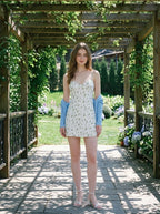 Woman in a white floral print mini dress and a blue cardigan standing in a garden.