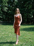 Woman standing in a rust-colored satin slip dress with spaghetti straps and a cowl neckline, in a grassy outdoor setting with trees in the background.
