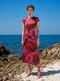 Woman wearing a pink and red floral print satin ruched midi dress standing on a rocky beach.