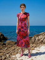 Woman wearing a pink and red floral print satin ruched midi dress standing on a rocky beach.