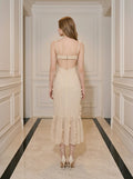 Back view of a woman wearing a cream-colored midi dress with 3D floral embellishments and a ruffled hem, standing in a hallway.