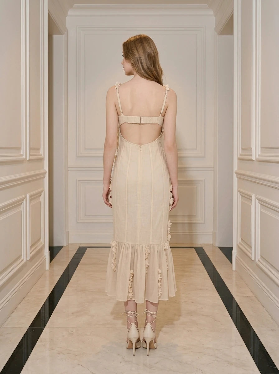 Back view of a woman wearing a cream-colored midi dress with 3D floral embellishments and a ruffled hem, standing in a hallway.