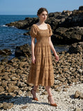 Woman walking on a rocky beach wearing a tan midi dress with a smocked bodice, puff sleeves, and a tiered skirt with a sun and Greek key print.
