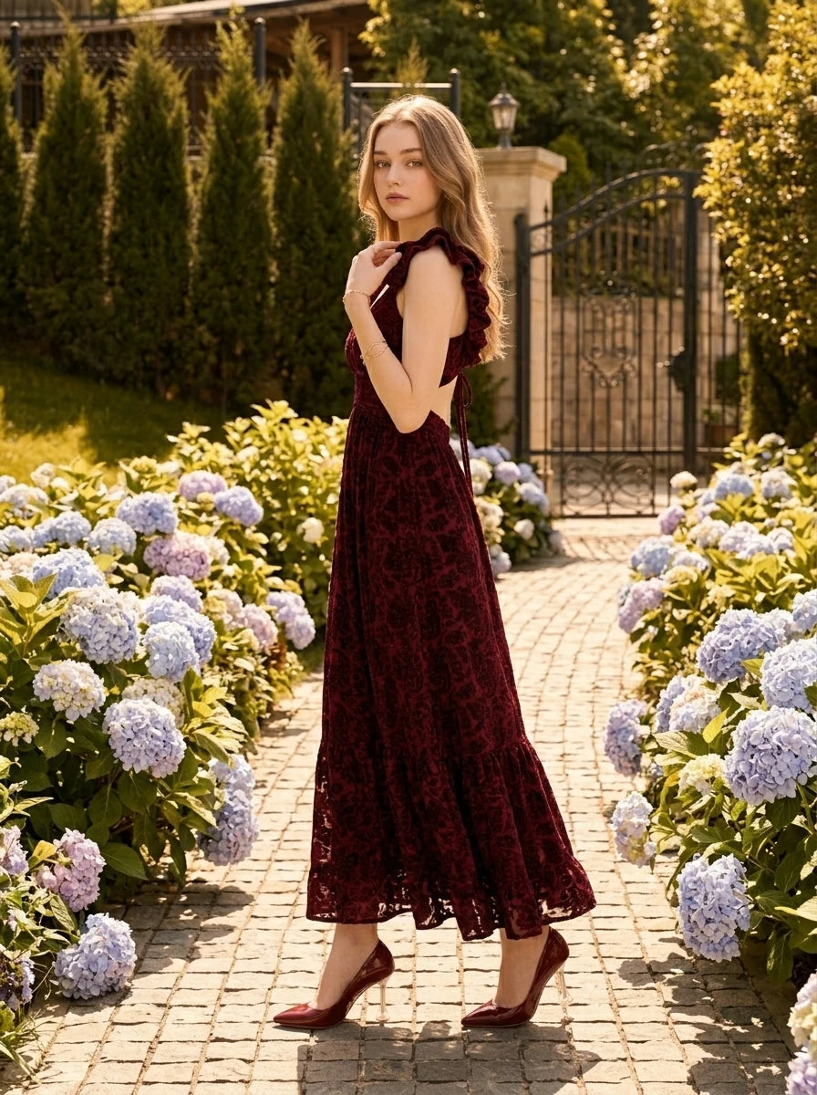 Woman in a burgundy velvet burnout A-line maxi dress with a V-neckline and ruffled cap sleeves, posing in a garden.