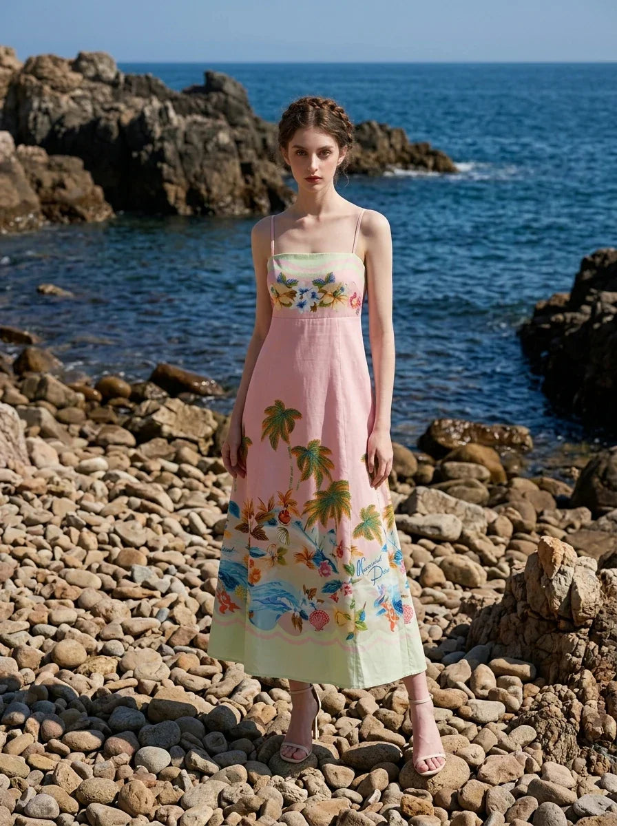 Woman standing on a rocky beach wearing a pink midi dress with spaghetti straps and a tropical print.