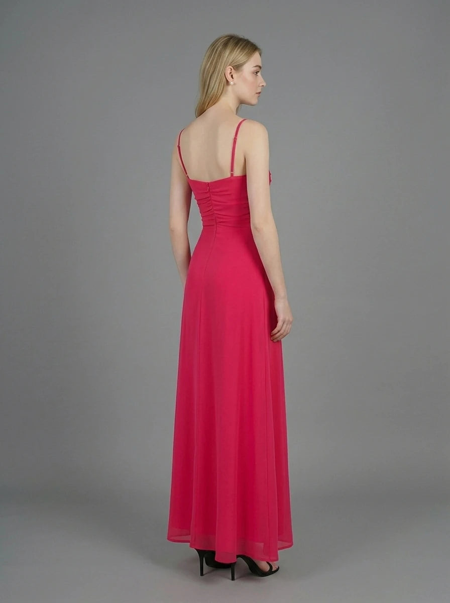 Woman in a bright pink ruched maxi dress with spaghetti straps and butterfly details, standing against a grey backdrop.