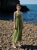 Woman wearing a green linen blend midi dress with a smocked bodice and spaghetti straps, standing on a pebble beach.