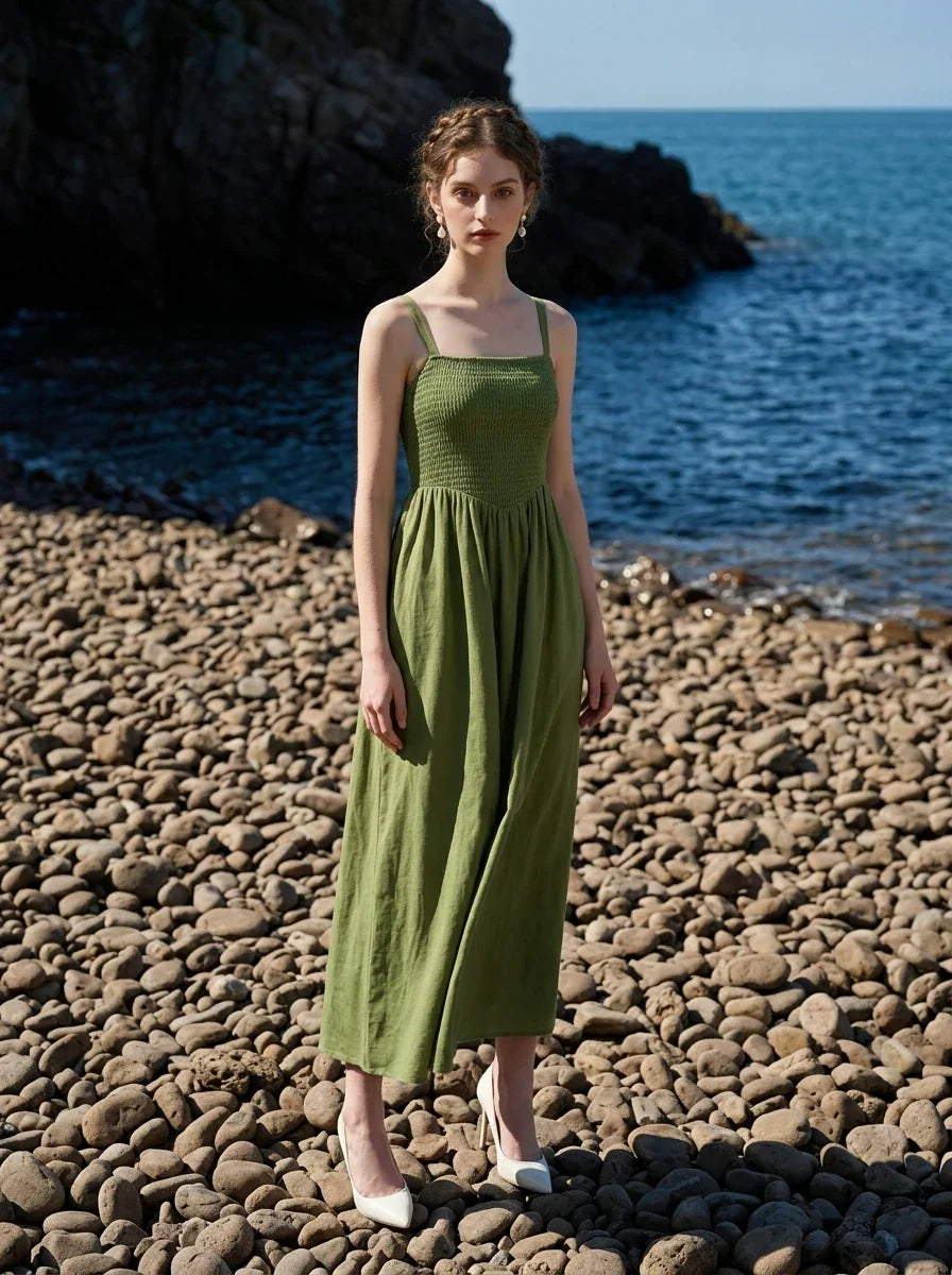 Woman wearing a green linen blend midi dress with a smocked bodice and spaghetti straps, standing on a pebble beach.