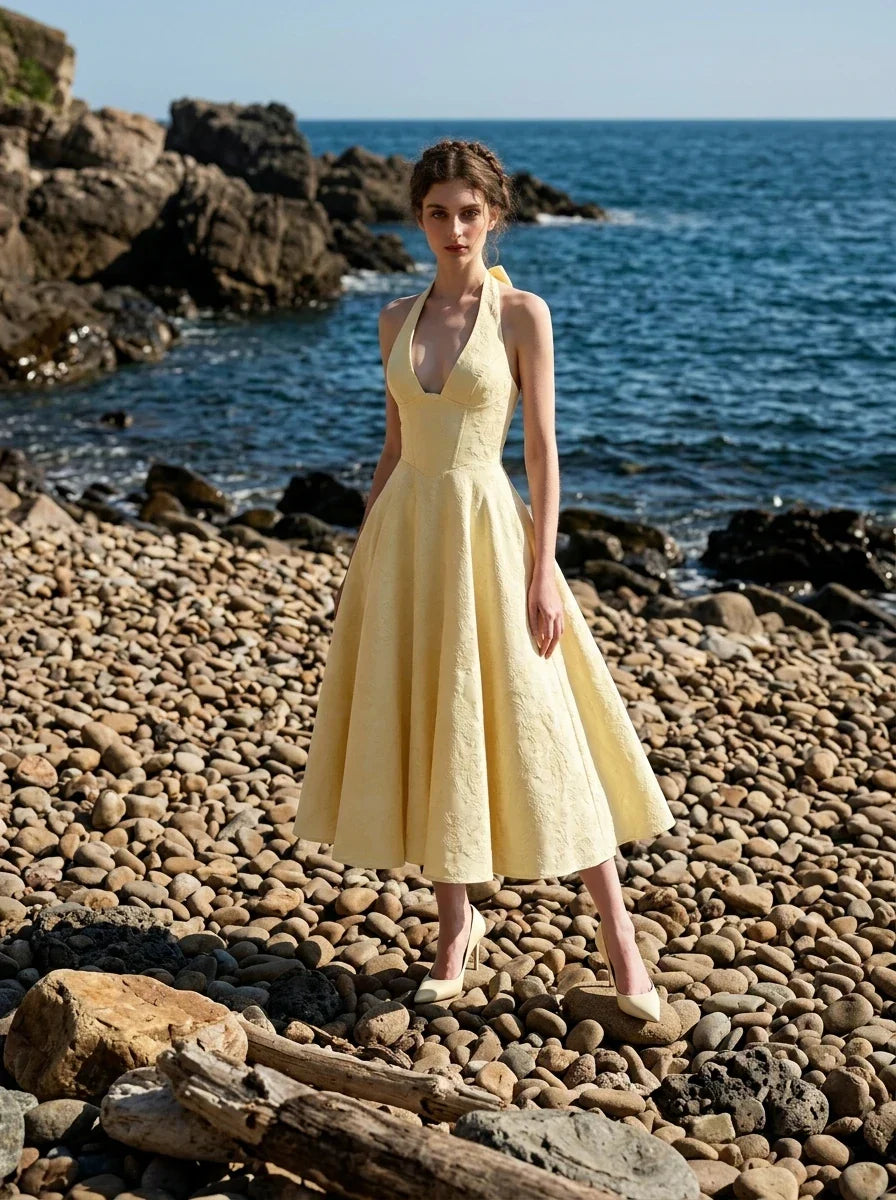 Woman in a pale yellow jacquard halter neck midi dress standing on a rocky beach.