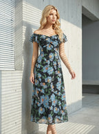 Woman standing outdoors wearing a black floral print off-the-shoulder midi dress with blue flowers, with shadows on the wall