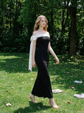Woman in a black off-the-shoulder mermaid dress with a white draped accent on the shoulder, walking in a grassy outdoor setting with trees.