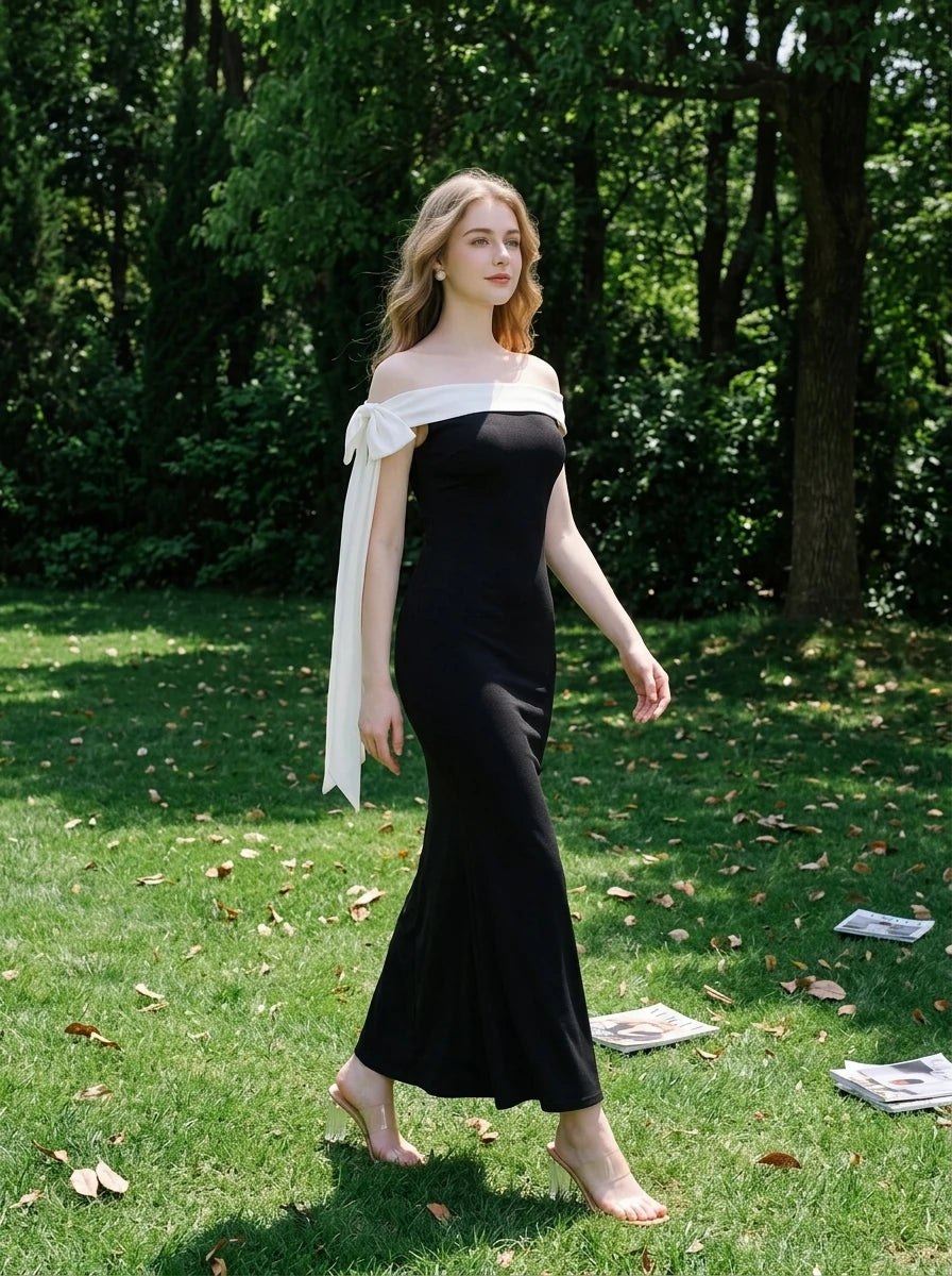 Woman in a black off-the-shoulder mermaid dress with a white draped accent on the shoulder, walking in a grassy outdoor setting with trees.