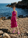 Woman wearing a fuchsia satin halter neck tiered midi dress with a self-tie belt, standing on a pebble beach by the ocean