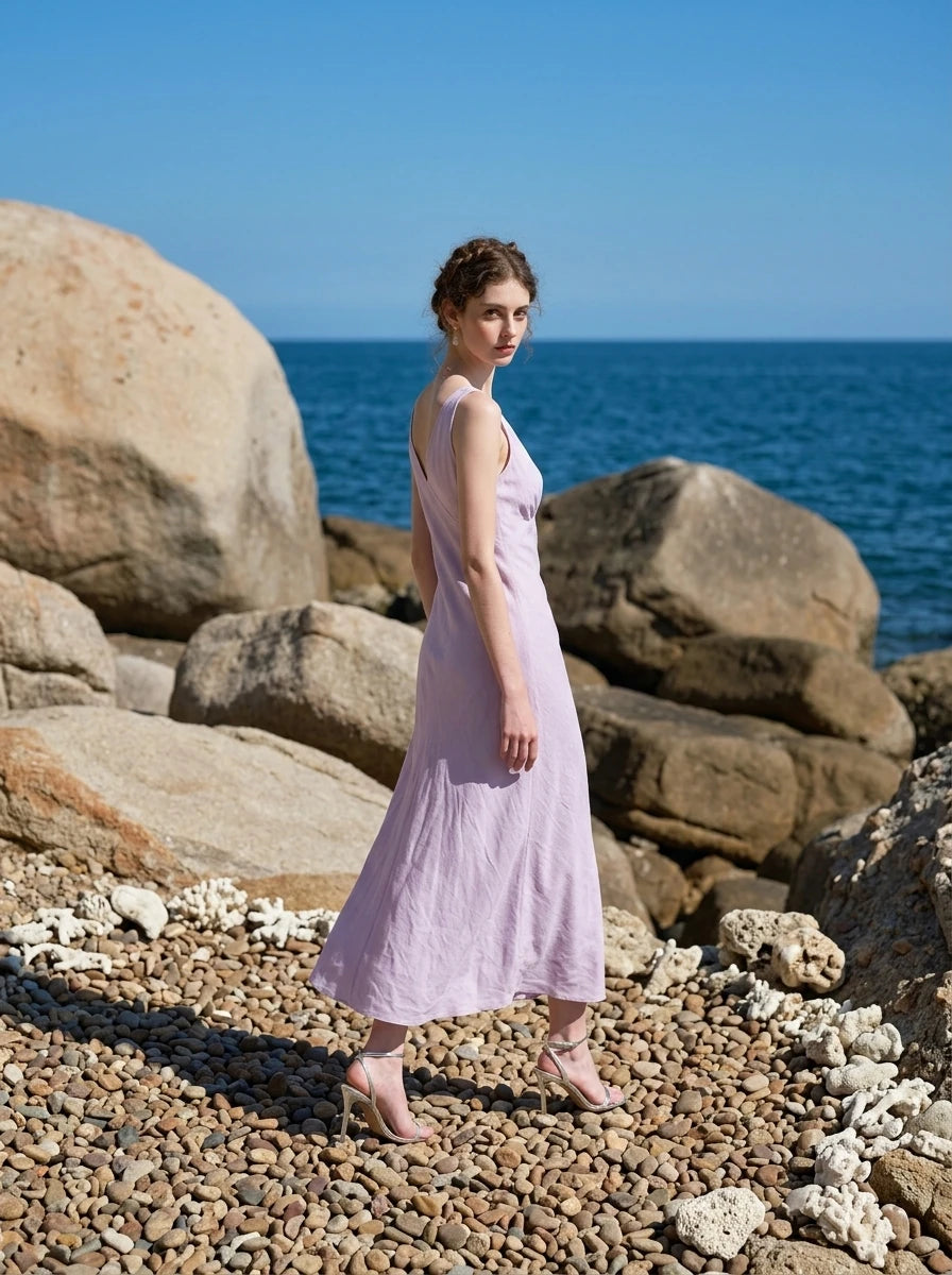 Woman in a lilac midi dress with a V-neckline and sleeveless design, walking on a rocky beach with the ocean behind her.