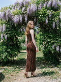Woman in a brown maxi dress standing in a garden with purple flowers.