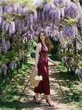 Woman in a burgundy lace and satin halter neck dress walking through a garden with wisteria