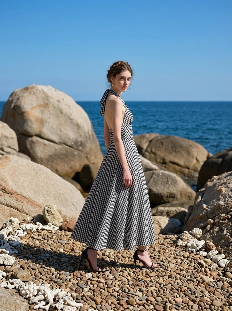 Side view of a woman wearing a black and white gingham check halter neck maxi dress with a plunging V-neck and a defined empire waist, posing on a beach with rocks and the ocean in the background.