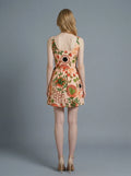 Back view of a woman wearing a sleeveless, scoop neck, fit and flare A-line dress with an all-over print of strawberries, kiwis, flowers, and coffee cups, in a peachy-pink hue.