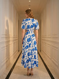 Back view of a woman wearing a white and blue floral print midi dress with puff sleeves and a cut-out waist, standing in a hallway.