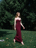 Woman wearing a floor-length, strapless burgundy gown with ruching and gold embellishment, walking in a grassy park.