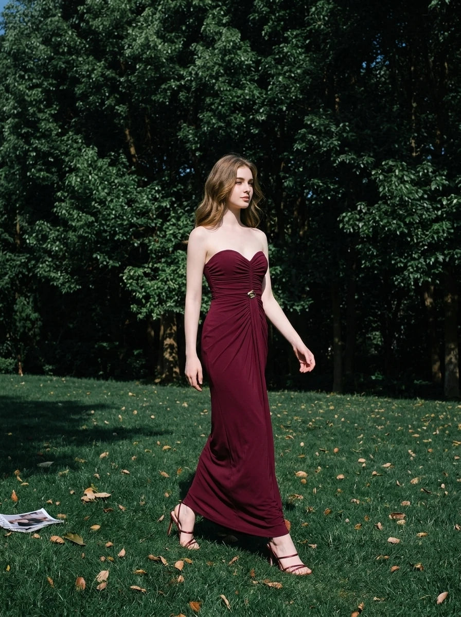Woman wearing a floor-length, strapless burgundy gown with ruching and gold embellishment, walking in a grassy park.