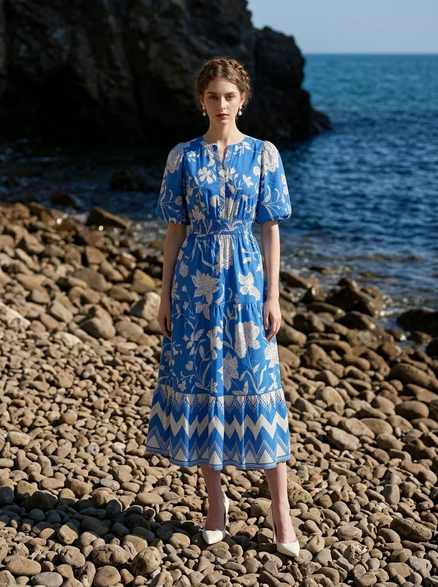 Woman wearing a blue floral print midi dress with puff sleeves, standing on a rocky beach with the ocean in the background.