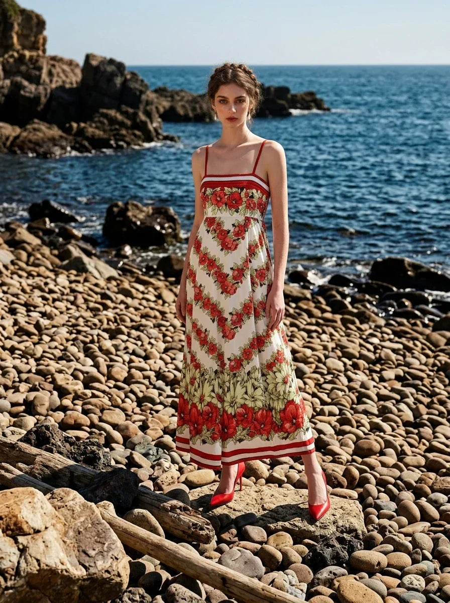 Woman wearing a white maxi dress with a red floral print and red trim, posing on a rocky beach with the ocean in the background.
