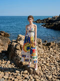 Woman standing on rocks by the sea wearing a white midi dress with a colorful abstract graphic print and gold chain spaghetti straps.
