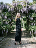 Woman in a black long-sleeved V-neck gown with a front knot and high slit, standing in a garden with purple wisteria.