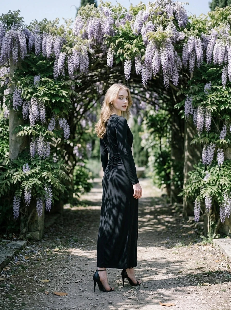 Woman in a black long-sleeved V-neck gown with a front knot and high slit, standing in a garden with purple wisteria.
