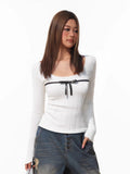 Close-up of a woman's upper body, showcasing a white ribbed long-sleeve top with a square neckline and a black ribbon bow.