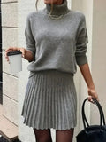 Woman in a grey knit turtleneck sweater and pleated mini skirt holding a coffee cup and a black handbag.