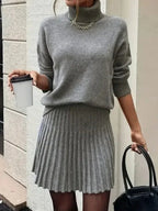 Woman in a grey knit turtleneck sweater and pleated mini skirt holding a coffee cup and a black handbag.