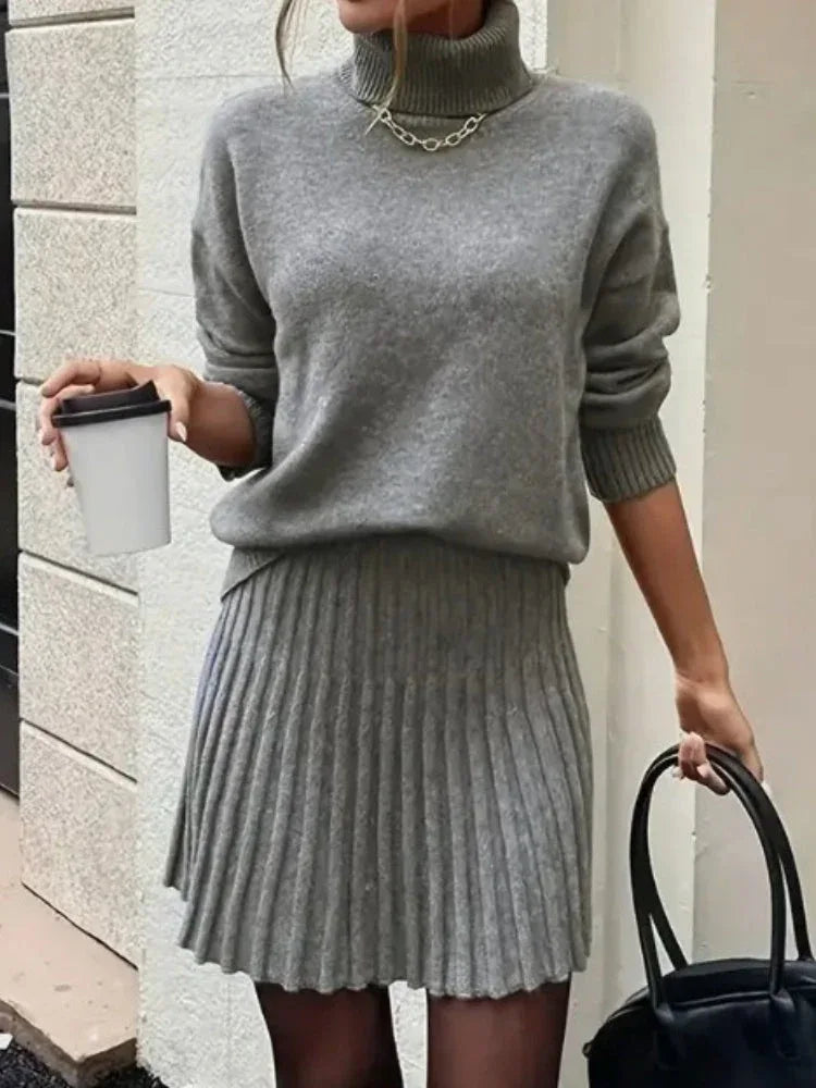 Woman in a grey knit turtleneck sweater and pleated mini skirt holding a coffee cup and a black handbag.