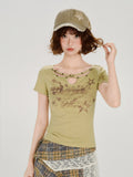 Woman wearing a light green short-sleeve fitted t-shirt with a cut-out neckline detail featuring star embellishments and a central ring. The t-shirt has a graphic print on the front and is paired with a plaid skirt.