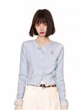 Woman wearing a light blue knit cardigan with a flower embellishment and white pants.