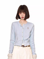 Woman wearing a light blue knit cardigan with a flower embellishment and white pants.