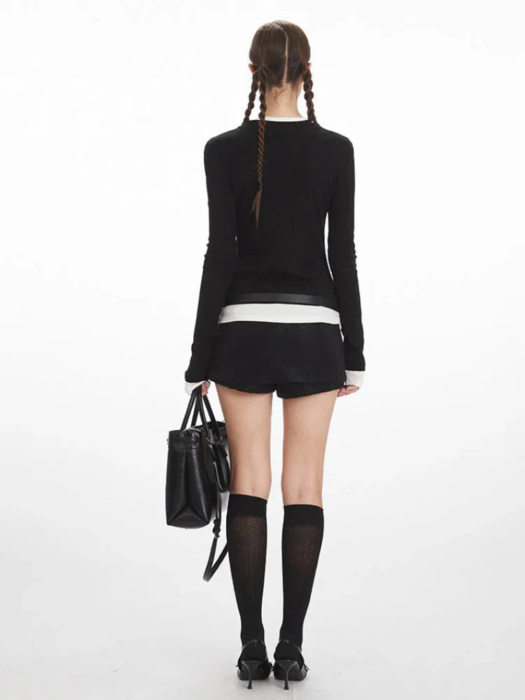 Back view of a model in a black long-sleeve top with white contrast trim, layered over a white band, paired with black shorts and knee-high socks.