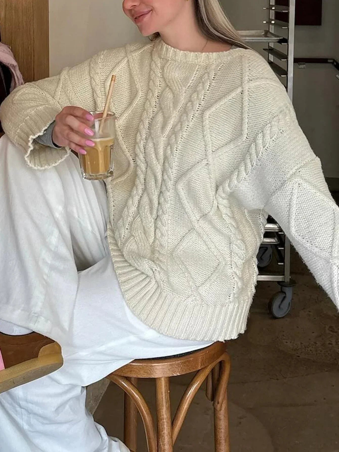 Woman wearing a cream cable knit sweater and white pants, holding a coffee drink. Main image