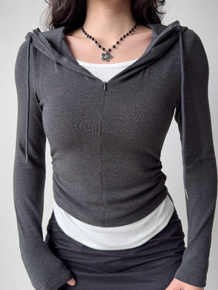 Close-up of a woman wearing a charcoal grey ribbed knit zip-up hoodie with a drawstring hood and ruched sides, layered over a white tank top.