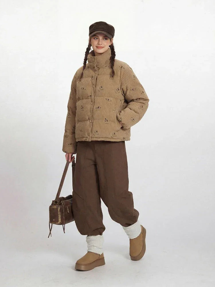 Full body shot of a woman in a tan corduroy puffer jacket with a dog print and brown pants.